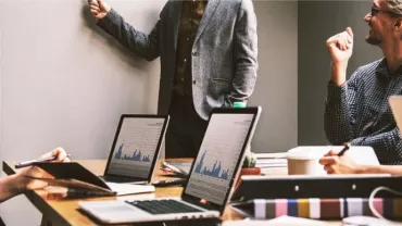 Riunione in ufficio con persone che discutono e laptop aperti sul tavolo.